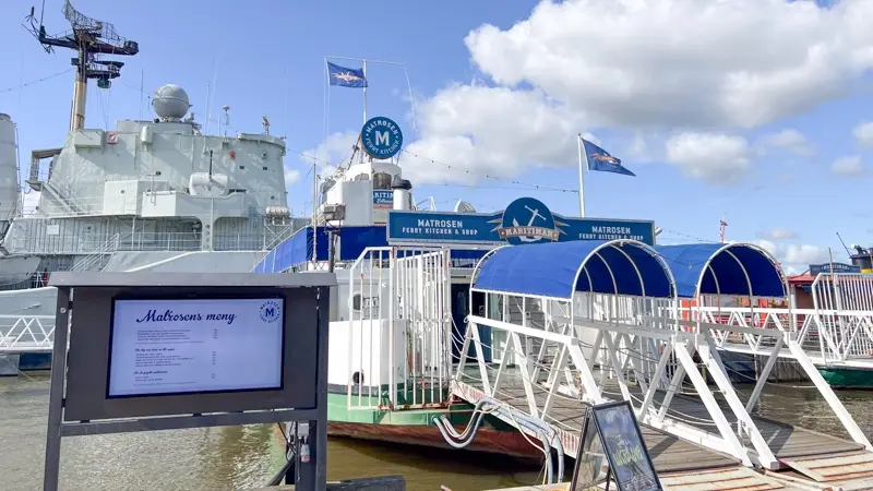 Outdoor digital signage display by a waterfront with a restaurant and ferry pier in the background.