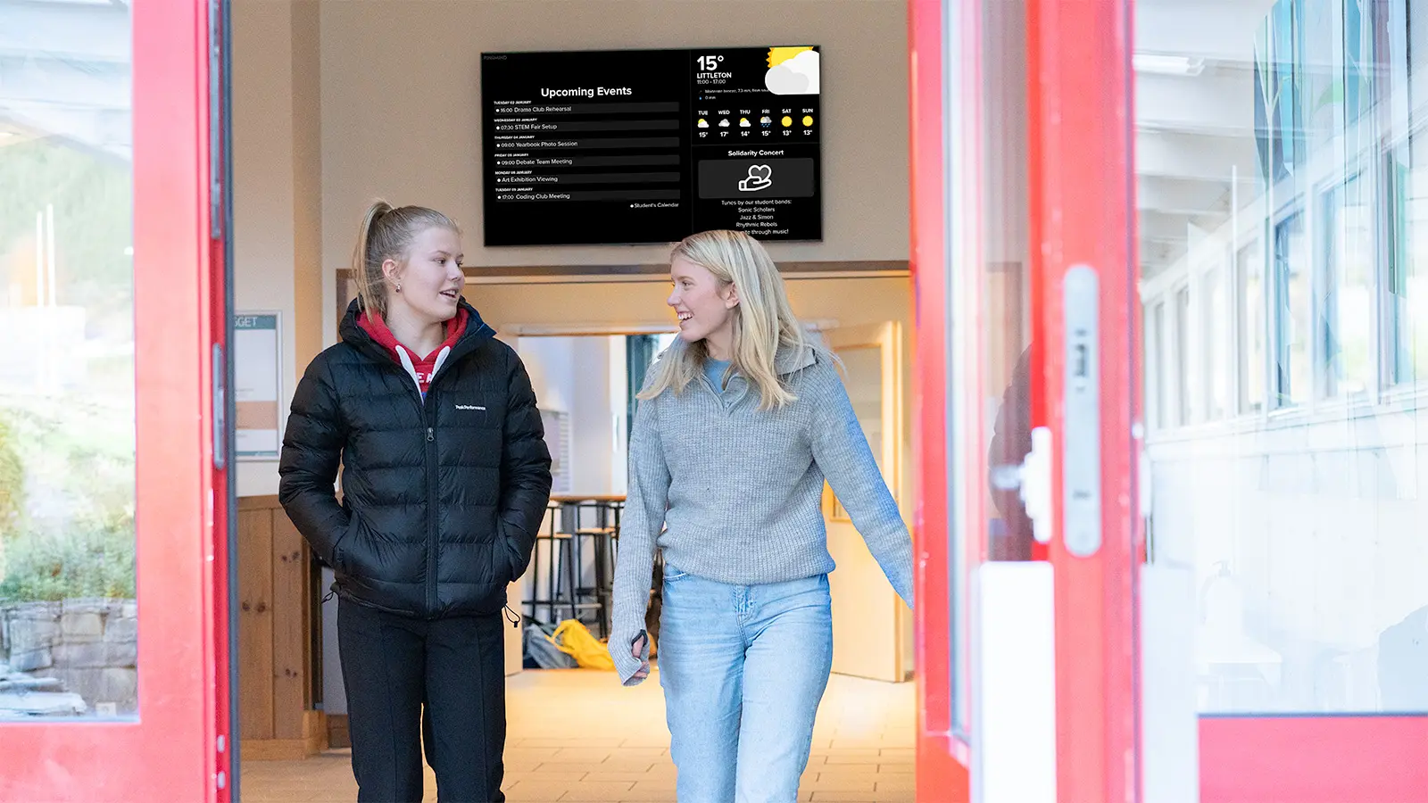 Two students leaving a high school, with a digital display visible in the background.
