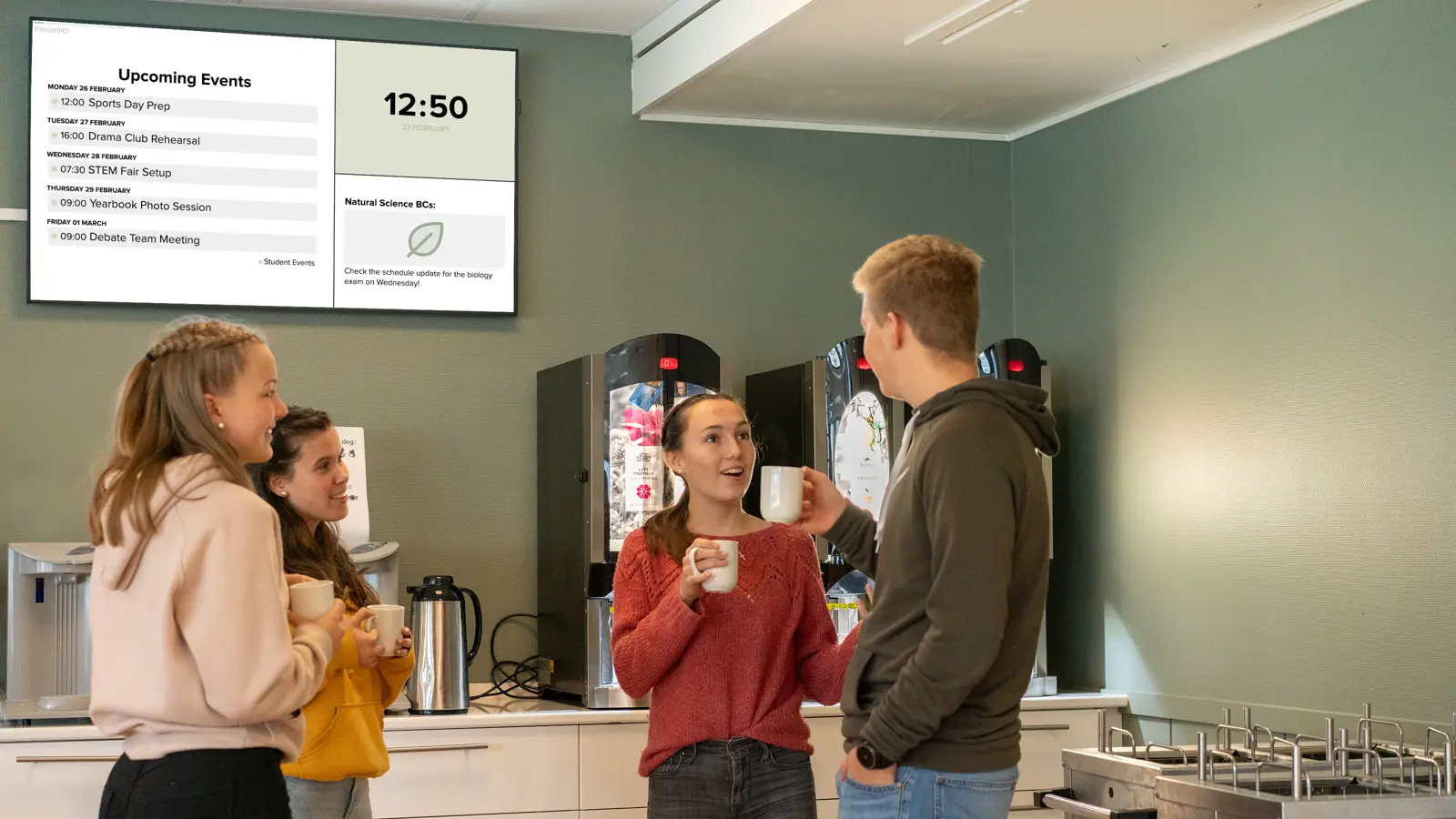Three students talking next to a digital display in the school canteen.